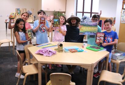 A group of children and an adult displaying board games around a table in a classroom setting.