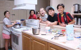 Six kids gather in a kitchen while one cooks on the stove. Baking ingredients and utensils are on the counter.