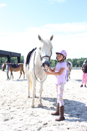 A girl in riding gear stands beside a white horse on a sunny day. Other riders and horses are in the background.