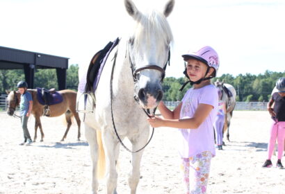 A girl in riding gear stands beside a white horse on a sunny day. Other riders and horses are in the background.