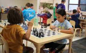 Children playing chess in a classroom setting, seated at tables with chessboards. Volunteers are present, assisting the children.