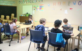 A classroom with children seated at desks using laptops and tablets, with colorful letters spelling "HOMEWORK" on the wall.