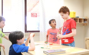 Four boys are gathered around a classroom table, engaged in a lively discussion or game. One boy stands, gesturing, while another looks surprised. A "Fire Exit" sign is visible on a window.