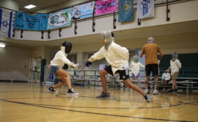 Two people fencing in a gym, wearing protective gear. Other participants and a coach are in the background.