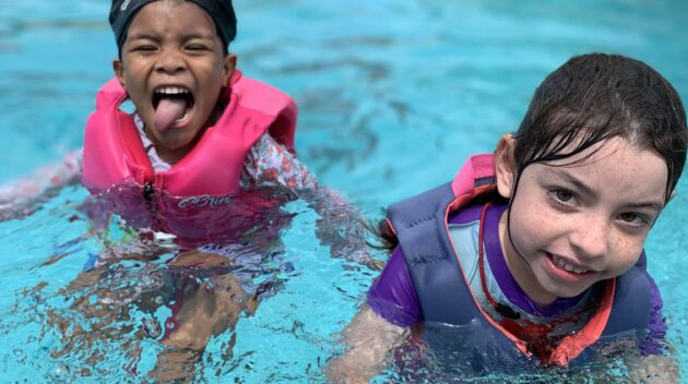 Two children in pink and purple life vests playing in a swimming pool, one making a playful face.