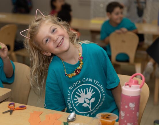 Child with blonde hair wearing a teal shirt and cat ear headband, sitting at a table with craft materials and a pink water bottle. Other children and chairs are in the background.