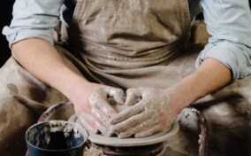 Person shaping clay on a pottery wheel, hands covered in wet clay, wearing a dirty apron and light blue shirt.