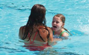 An adult and a child in swimwear play in a pool. The child has her tongue out, while the adult faces her. The sun reflects off the water.