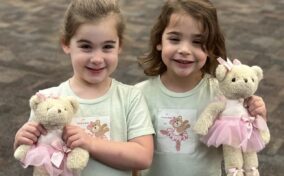 Two young girls smiling and holding teddy bears dressed in pink tutus. Both girls are wearing light green shirts with bear graphics and standing on a carpeted floor.