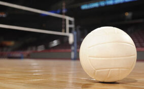 A volleyball rests on a polished indoor court with a net in the background.