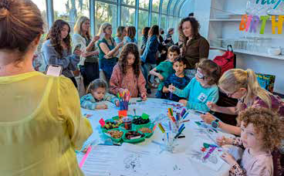 Children and adults gather around a table with art supplies, coloring and drawing, in a bright indoor space.