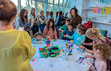 Children and adults gather around a table with art supplies, coloring and drawing, in a bright indoor space.