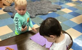 Two toddlers at a table, one holding a toy elephant and the other looking down at papers. A toy giraffe is on the table.