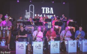 A big band, including saxophonists and brass players, performs on stage. A screen behind them displays "TBA Big Band." Musicians are seated with stands and instruments.