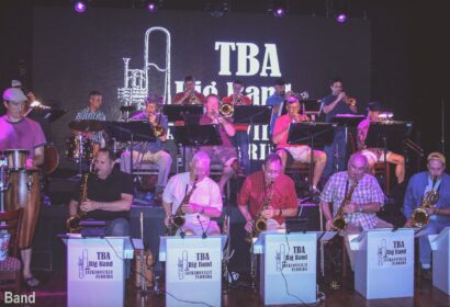 A big band, including saxophonists and brass players, performs on stage. A screen behind them displays "TBA Big Band." Musicians are seated with stands and instruments.