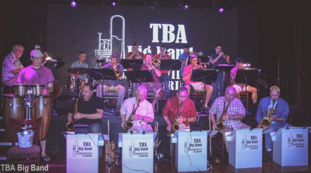 A big band, including saxophonists and brass players, performs on stage. A screen behind them displays "TBA Big Band." Musicians are seated with stands and instruments.