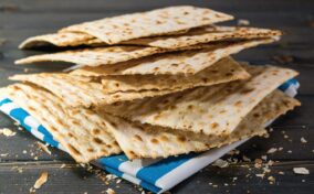 A stack of rectangular matzah crackers on a wooden surface with a blue and white cloth underneath. Crumbs are scattered around.