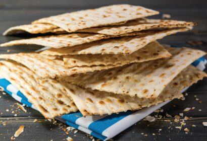 A stack of rectangular matzah crackers on a wooden surface with a blue and white cloth underneath. Crumbs are scattered around.