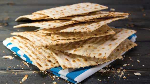 A stack of rectangular matzah crackers on a wooden surface with a blue and white cloth underneath. Crumbs are scattered around.