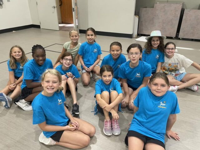 A group of children in blue shirts sitting on the floor of a room, smiling at the camera.