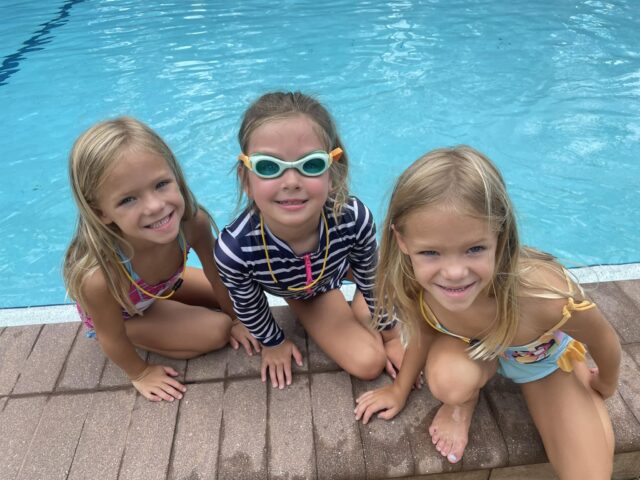 Three children in swimsuits sit by the edge of a swimming pool. One child wears goggles and a striped shirt.