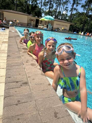 Five children in swimsuits and goggles sit at the edge of a pool, smiling. Trees and a building are visible in the background.