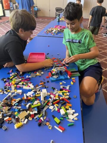 Two boys are building with colorful LEGO bricks on a blue table in a room with a patterned carpet.