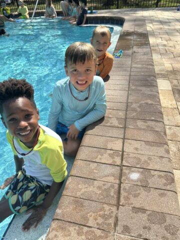 Three smiling children sit by the edge of a swimming pool, wearing swimwear and necklaces.