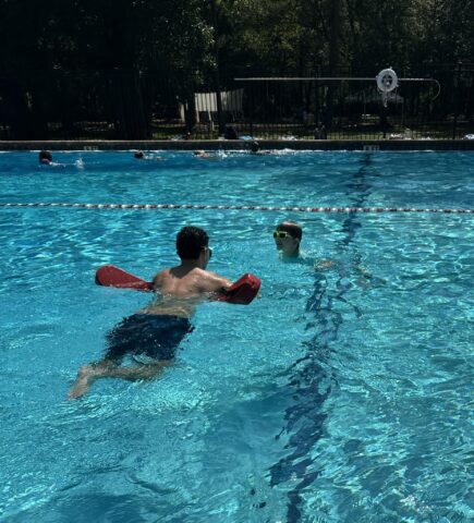 Two people are swimming in an outdoor pool. One person wears red swim aids and the other wears goggles. The water is clear, and several people swim in the background. Trees line the pool's edge.