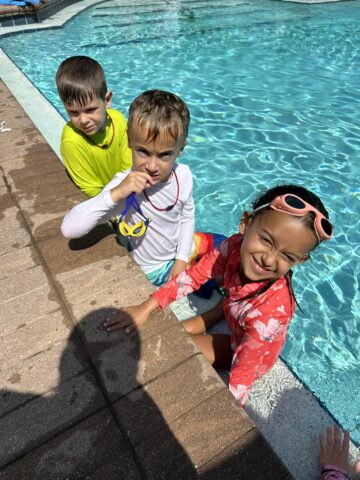 Three children in swimwear are at the edge of a pool. Two boys are in the water, one in yellow and one in blue. A girl in red is next to them, smiling, with pink goggles on her head.