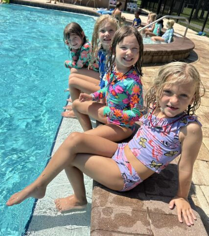 Four children in colorful swimsuits sitting poolside with their feet in the water, smiling on a sunny day.