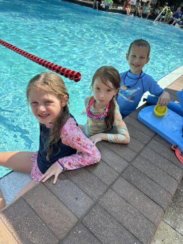 Three children sit by a pool, wearing swimwear. Two girls are in the water while a boy sits on the edge with swimming gear beside him.