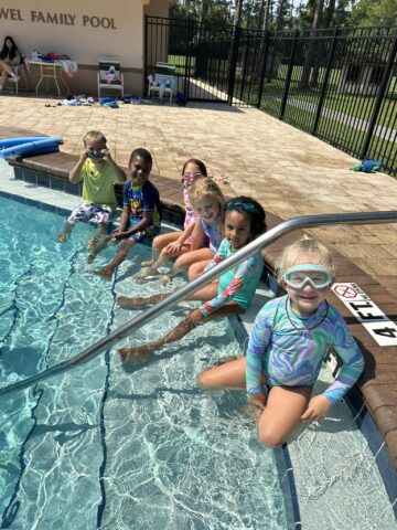 A group of children sitting on the edge of a pool, wearing swimwear and goggles, with their feet in the water.