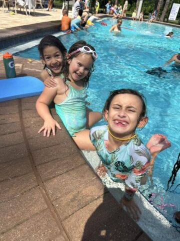 Three children at the edge of a swimming pool, smiling and wearing swimwear. Others swim in the background.