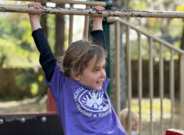 A child in a purple shirt hangs from playground monkey bars, smiling, with trees and playground equipment in the background.