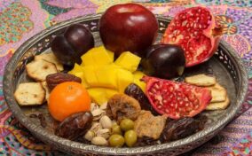 A decorative tray with pomegranate, apple, plums, mango pieces, crackers, tangerine, dates, olives, pistachios, and dried figs on a colorful tablecloth.