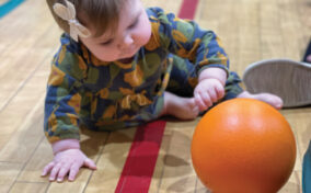 A baby on a gym floor reaches for an orange ball, wearing a patterned outfit with a small bow in their hair.