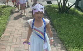 A child in a costume with a butterfly mask walks on a brick path outside. Other children follow behind, and trees line the path.