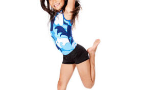 A young girl wearing a blue and white leotard jumps energetically against a white background, smiling with arms raised and one leg bent backward.