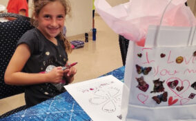 Young girl smiling at a table with art supplies, next to a decorated gift bag. Children are seen in the background.