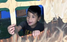 Child looking out of a painted cardboard cutout window, appearing engaged and cheerful.