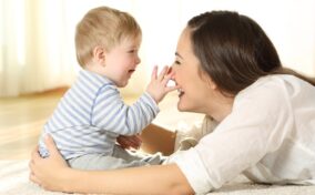 A baby wearing a striped shirt playfully touches a smiling woman's nose as they lie on a carpet.