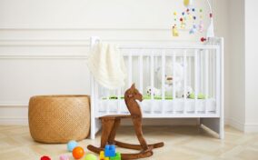 A white crib with toys and a teddy bear. A wicker basket is on the left, and a wooden rocking horse is in the foreground. Colorful blocks and balls are scattered on the floor.