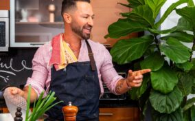 Person with a beard and apron smiling and gesturing in a kitchen, surrounded by greens and spices. Large plant visible in the background.