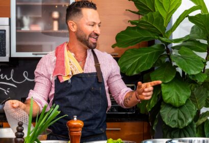 Person with a beard and apron smiling and gesturing in a kitchen, surrounded by greens and spices. Large plant visible in the background.