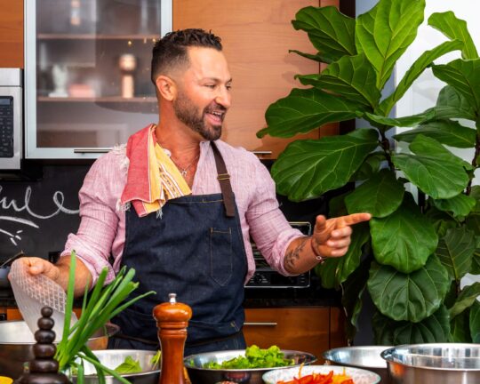 Person with a beard and apron smiling and gesturing in a kitchen, surrounded by greens and spices. Large plant visible in the background.