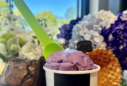 A cup of purple ice cream with a green spoon, topped with a cookie piece. A brownie and a waffle are placed beside it. Flowers and a window view are in the background.