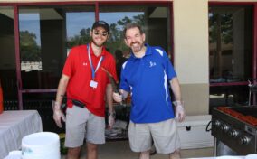 Two men stand by a grill cooking hot dogs, one holding tongs. They are smiling, wearing sunglasses and casual attire. Plates are stacked on a nearby table.