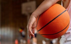 Person holding an orange basketball in a gym with a blurred hoop in the background.