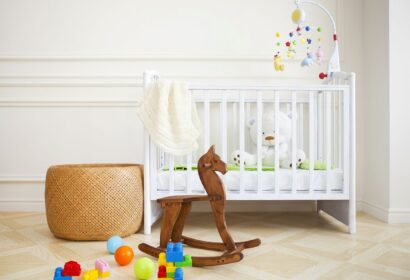 White crib with toys and mobile in nursery. Wooden rocking horse, balls, and building blocks on the floor. Beige woven basket nearby.